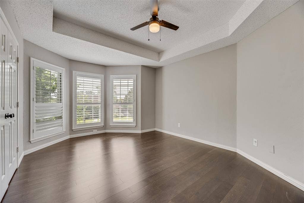 Recessed ceiling with a ceiling fan, bay window with plantation shutters, light gray wall paint, and dark wood-finish flooring