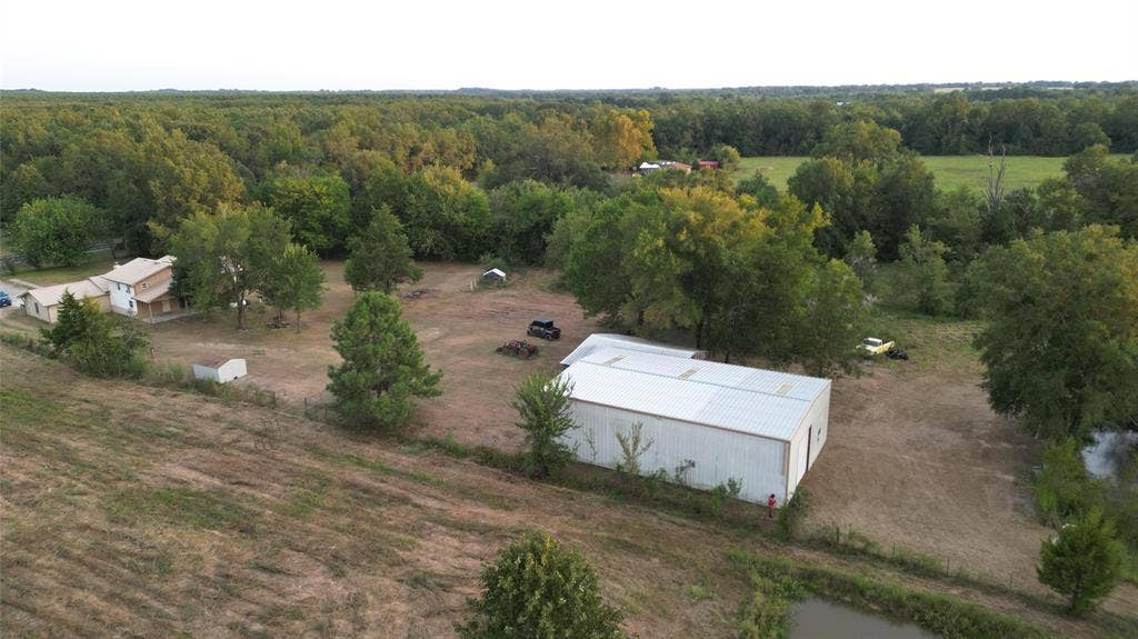 Expansive acreage featuring a residential structure, a detached metal workshop, multiple outbuildings, mature tree clusters, and a pond