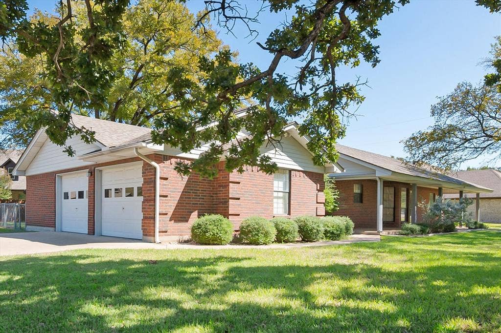 5203 Thunderbird Trail, De Cordova, TX 76049 Brick exterior with white siding accents, two-car garage with panel doors, covered front porch, and mature trees providing shade