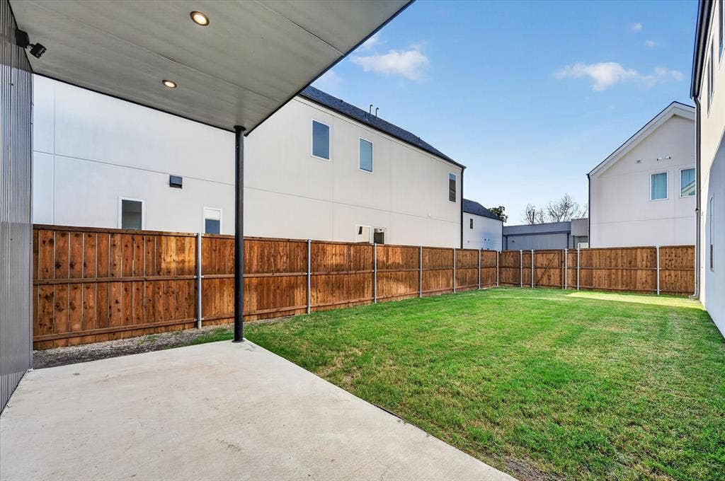 Expansive backyard featuring a manicured lawn, full perimeter wood fencing, and a concrete patio with an overhead cover and recessed lighting