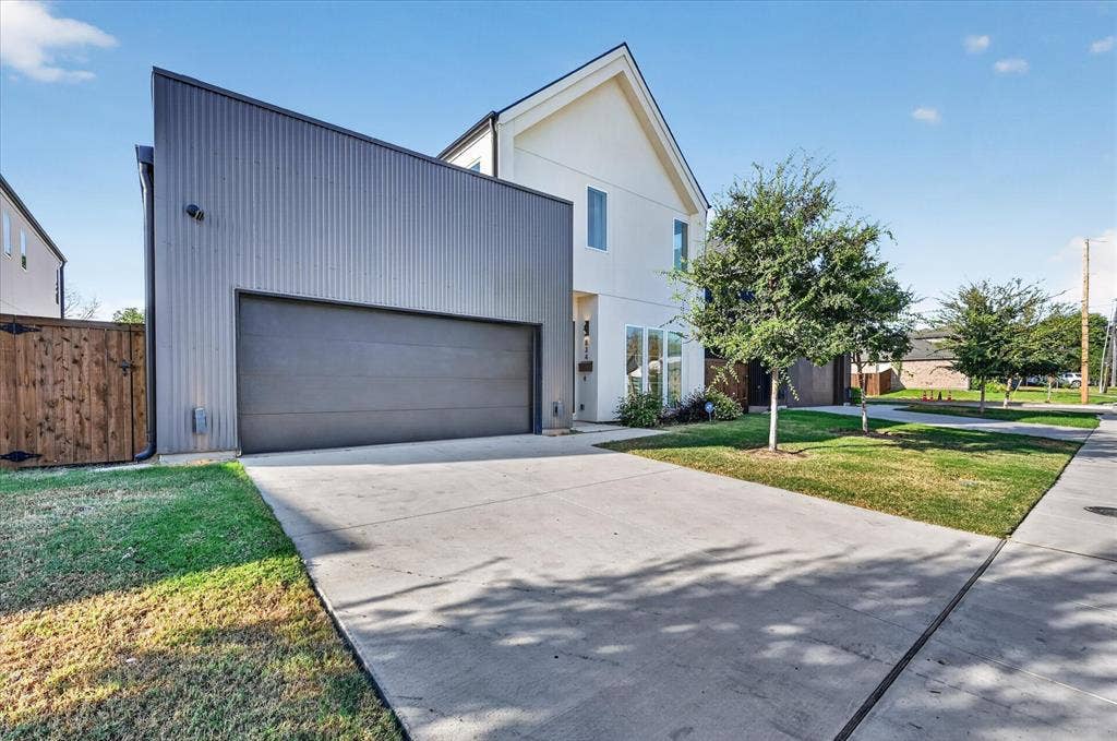 Contemporary exterior with a white facade and a gray corrugated metal accent, featuring a two-car garage with a gray door, a concrete driveway, and a manicured lawn with mature trees