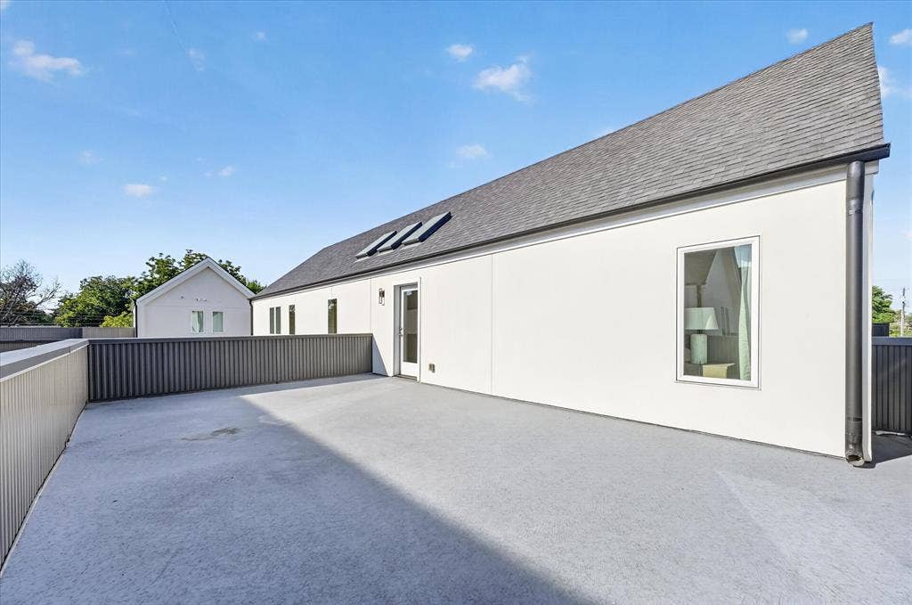 Spacious rooftop patio with concrete surfacing and contemporary slatted railings, featuring a white stucco exterior and dark shingle roofing