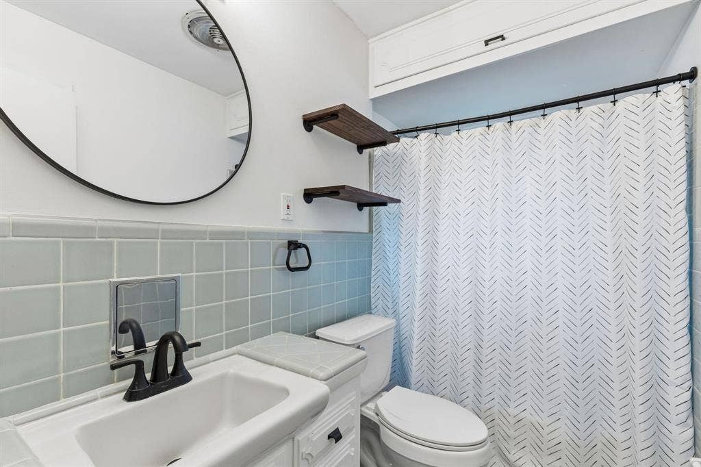 7230 Edgerton Drive, Dallas, TX 75231 Bathroom featuring light blue square tile backsplash, white vanity with integrated sink, matte black faucet, wall-mounted circular mirror, and built-in upper cabinetry