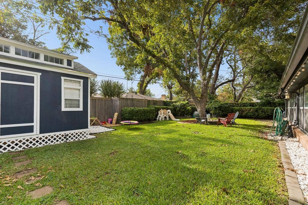 7230 Edgerton Drive, Dallas, TX 75231 Backyard featuring a manicured lawn, perimeter wood fencing, mature trees, a detached structure with transom windows, and white gravel landscaping accents