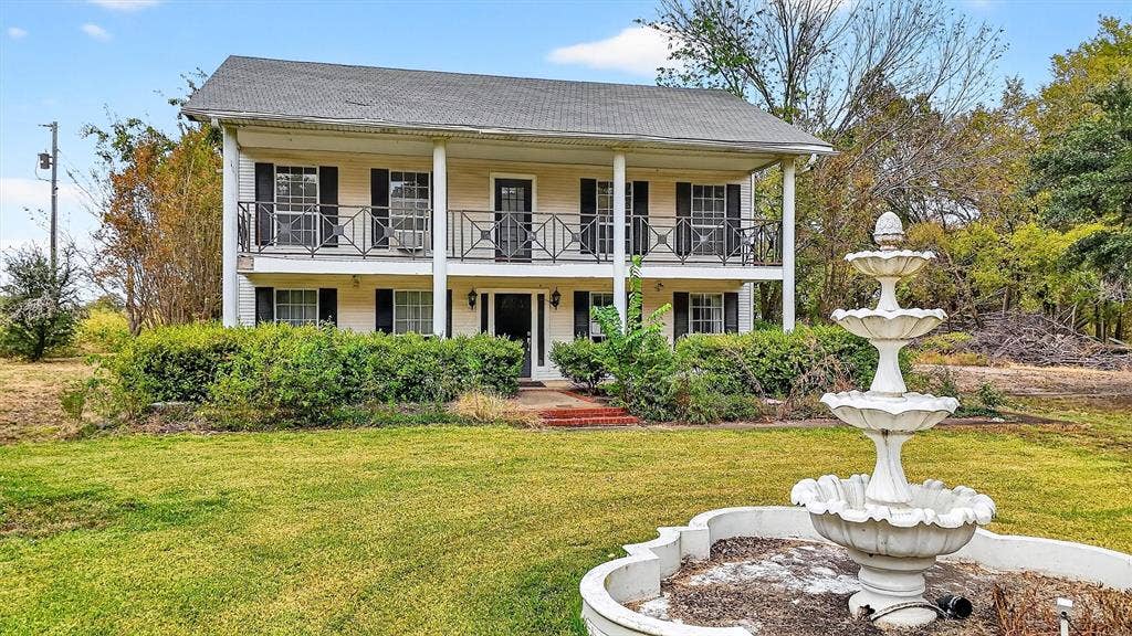 Two-story exterior with a full-width upper balcony, lower porch with columns, decorative iron railings, and a three-tiered fountain