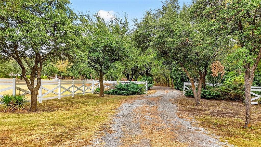Gravel driveway entrance with white crossbuck fencing, mature trees, and established shrubbery
