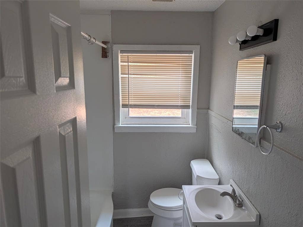 3409 Wood Avenue, Waco, TX 76711 Window with horizontal blinds, vanity with integrated sink, wall-mounted mirror, three-light vanity fixture, and toilet