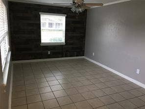 Room featuring neutral tone tiled flooring, a horizontal wood-finish accent wall, ceiling fan with light fixture, and a single window with blinds