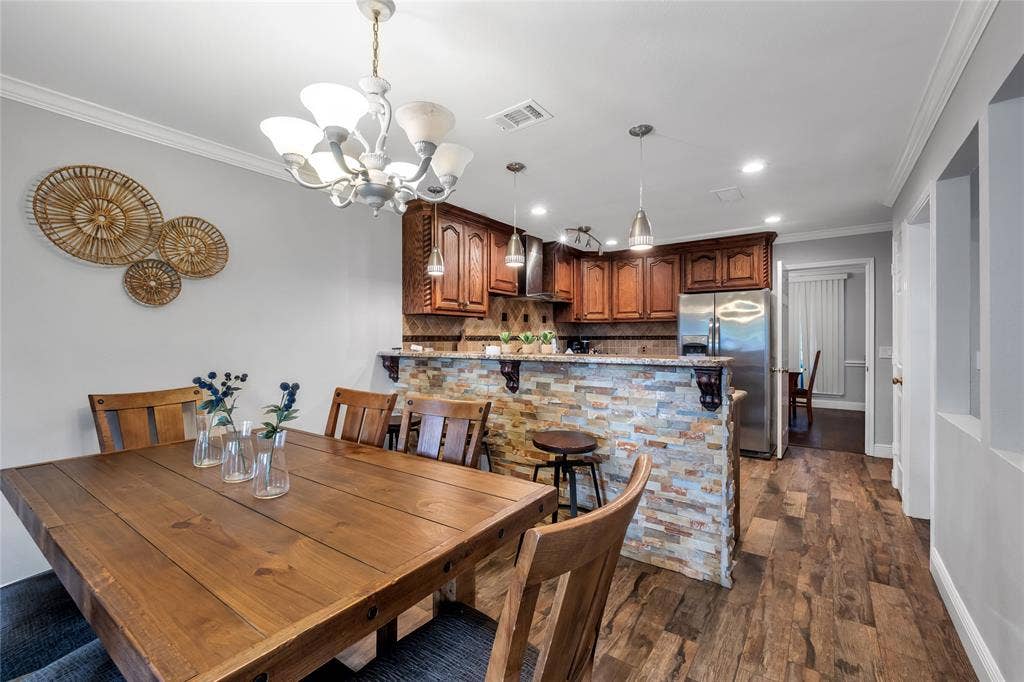 4523 Wescott Drive, Grand Prairie, TX 75052 Open-concept dining and kitchen area featuring warm wood cabinetry, stainless steel appliances, a multi-colored brick backsplash, wood-finish flooring throughout, and a decorative chandelier overhead