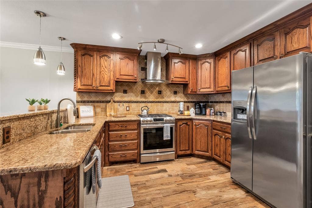 4523 Wescott Drive, Grand Prairie, TX 75052 Gourmet kitchen featuring rich wood cabinetry, granite countertops, stainless steel appliances including a side-by-side refrigerator and gas range, pendant lighting, and warm wood-finish flooring throughout the open layout