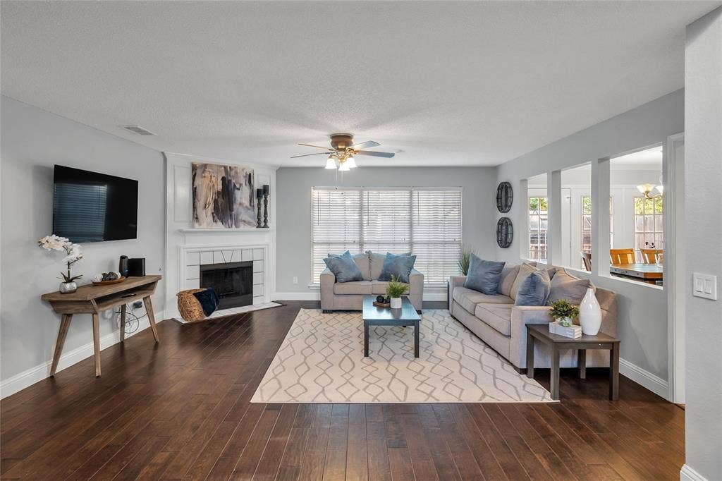 4523 Wescott Drive, Grand Prairie, TX 75052 Dark wood-finish flooring, white fireplace, multiple windows with blinds, ceiling fan, and a pass-through opening to an adjacent room