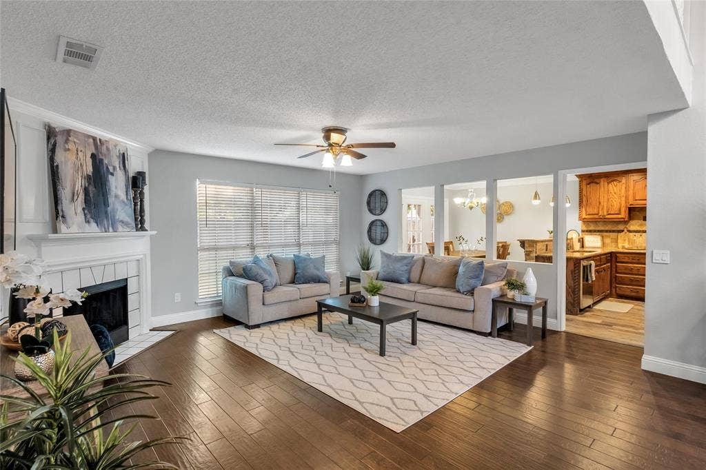 4523 Wescott Drive, Grand Prairie, TX 75052 Spacious living area featuring wood-finish flooring, a white mantel fireplace, large windows with blinds, a ceiling fan, and open doorways to an adjacent kitchen with wood cabinetry