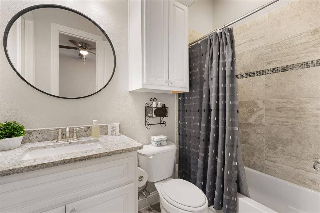 White vanity with a stone countertop and undermount sink, oval-framed mirror, white wall-mounted cabinet, and a tiled shower with an accent border