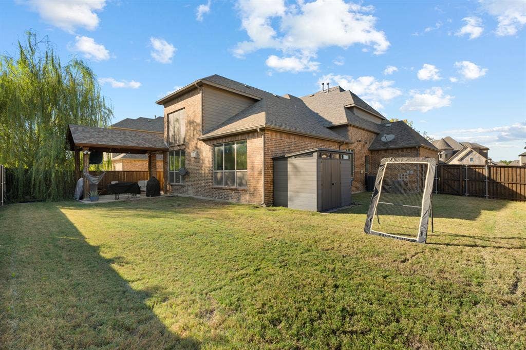 Expansive grass backyard featuring a covered patio with a wood-framed roof, brick exterior, and a permanent storage shed