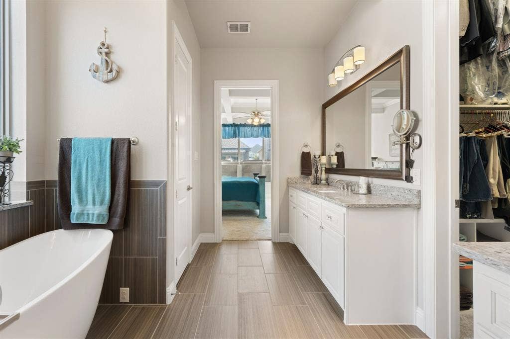 Freestanding soaking tub, wood-finish tile flooring, dual vanity with white cabinetry, framed mirror, and granite-style countertops