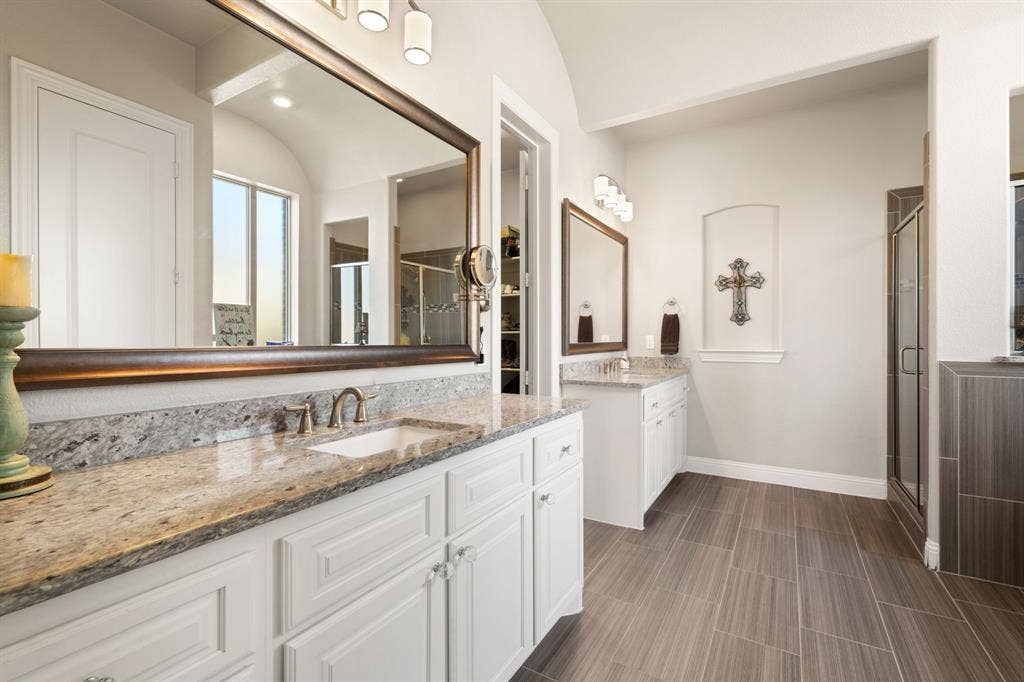 Dual vanity with stone countertops and undermount sinks, bronze-framed mirrors, wood-finish tile flooring, and a glass-enclosed shower