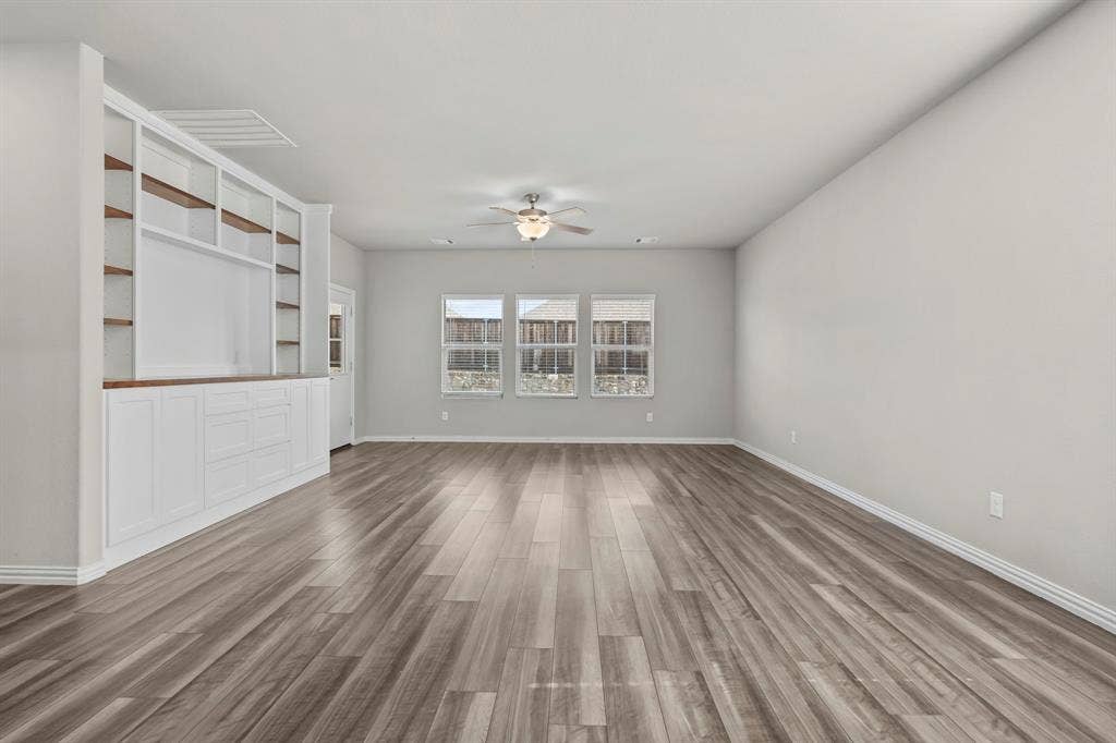 Expansive room featuring wood-finish flooring, three windows with white blinds, a ceiling fan with integrated lighting, and extensive built-in cabinetry with open shelving and closed storage