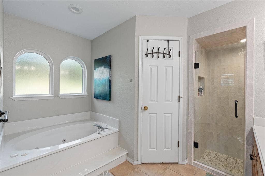 Jetted soaking tub with dual arched windows, new glass-enclosed shower with tile surround, and neutral tile flooring