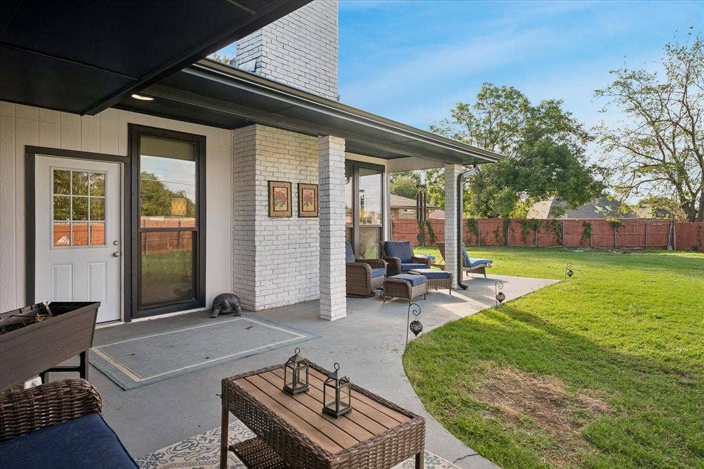 10102 Laingtree Drive, Dallas, TX 75243 Covered concrete patio featuring a white brick masonry pillar, exterior wall-mounted lighting, and a spacious backyard enclosed by wood fencing
