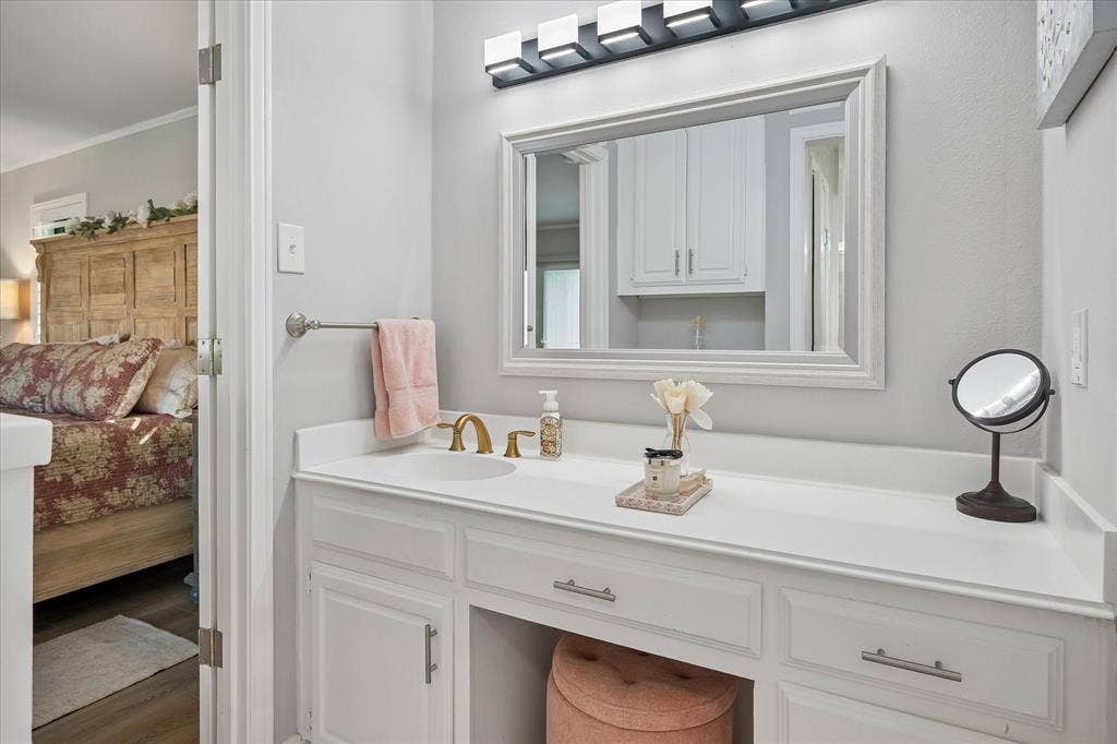10102 Laingtree Drive, Dallas, TX 75243 White built-in vanity featuring a solid surface countertop, integrated sink, brass-tone faucet hardware, and a framed mirror with integrated four-light fixture