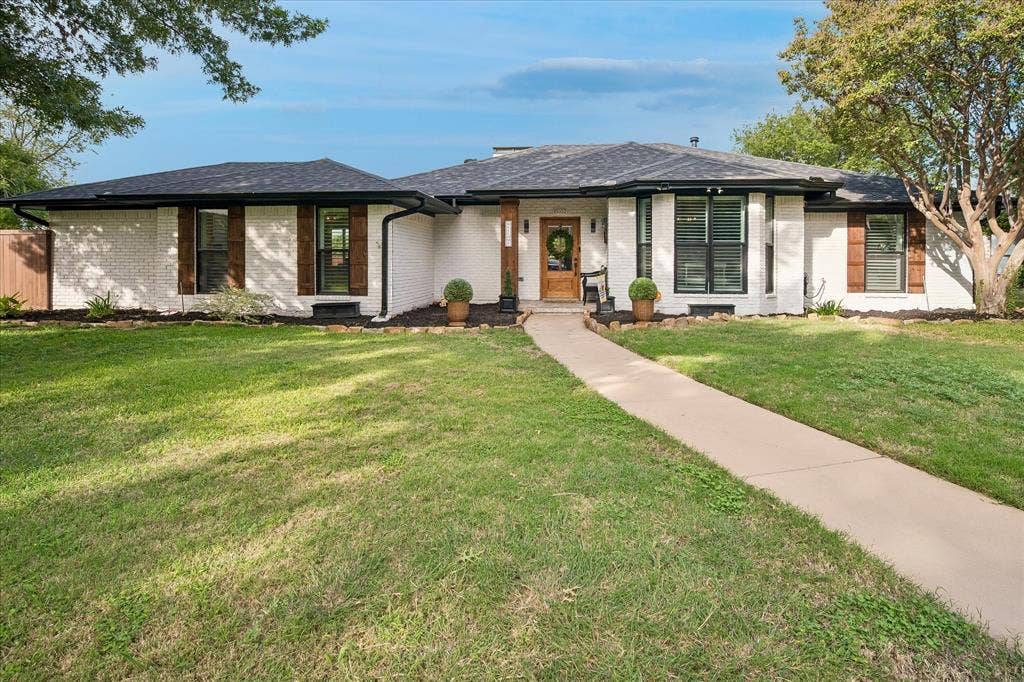 10102 Laingtree Drive, Dallas, TX 75243 Single-story exterior featuring painted brick masonry, dark-finish roof shingles, decorative wood-finish shutters, a covered entry with a timber-style column, and a concrete walkway
