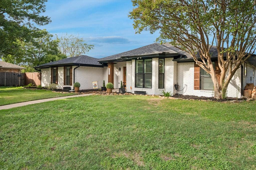10102 Laingtree Drive, Dallas, TX 75243 Single-story residence featuring white brick exterior, dark window trim, dark shingle roof, wood-tone column accent, and manicured lawn