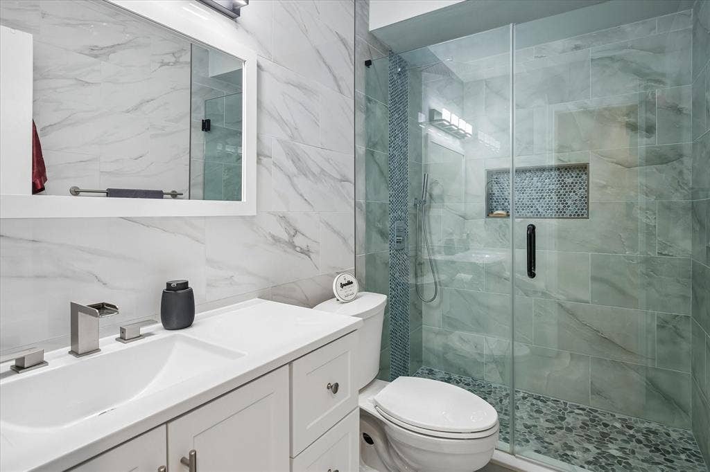 10102 Laingtree Drive, Dallas, TX 75243 Bathroom featuring a white vanity cabinet with an integrated sink, wall-mounted mirror, glass-enclosed walk-in shower with stone tile flooring, and a built-in recessed wall niche