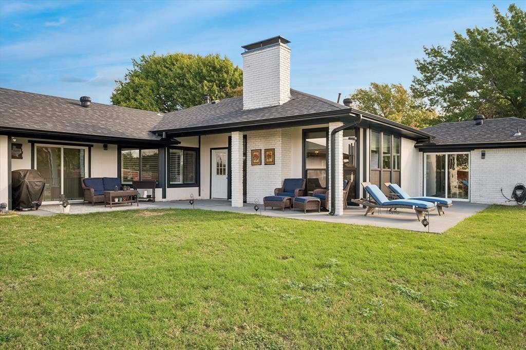 10102 Laingtree Drive, Dallas, TX 75243 Painted brick exterior featuring a central chimney, concrete patio, multi-pane windows, and sliding glass entry points
