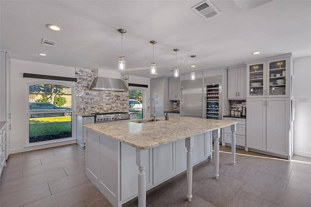 6802 Duffield Drive, Dallas, TX 75248 Kitchen featuring a central island with a light-toned countertop, stainless steel appliances, extensive cabinetry with some glass-front doors, recessed lighting, and a patterned tile backsplash