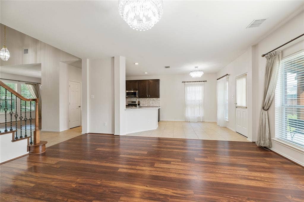 Wood-finish flooring, staircase with wrought iron balusters, white tiled flooring, recessed kitchen with dark wood-finish cabinetry, and multiple windows with blinds