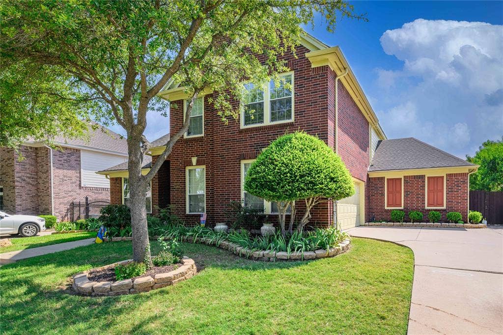 Red brick exterior with light trim, green lawn, mature tree, stone-edged flower bed, and a paved driveway