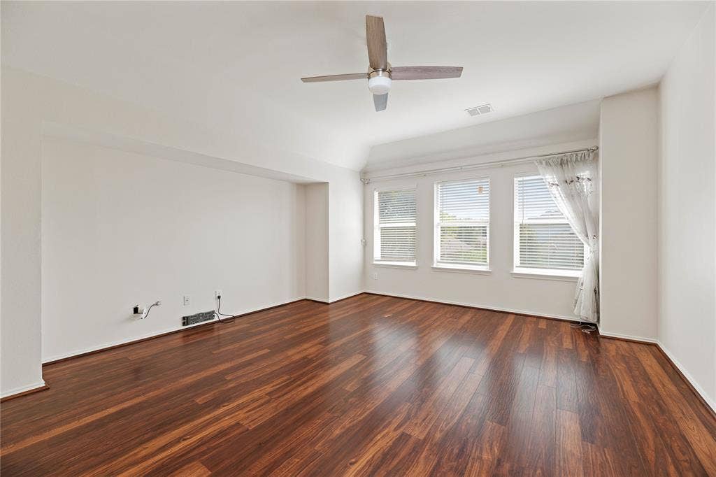 Spacious room showcasing rich wood-finish flooring, abundant natural light from multiple windows, ceiling fan with brushed metal finish, and fresh white walls with clean architectural lines