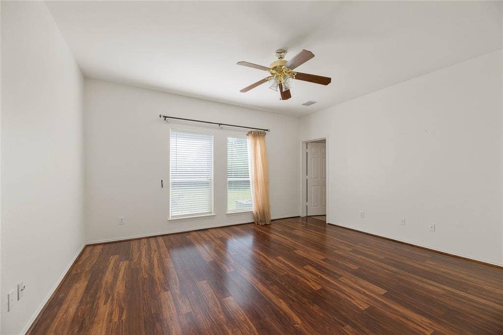 Spacious room flooded with natural light through dual glass doors and windows, featuring rich wood-finish flooring throughout and a ceiling fan with brass accents