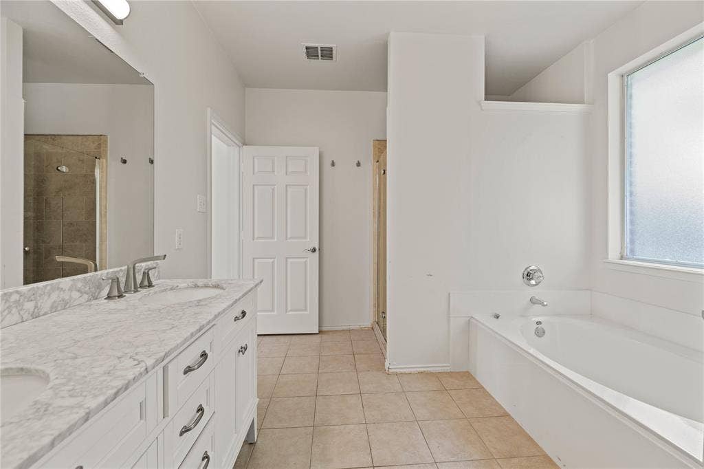 Spa-inspired primary bathroom featuring a dual-sink vanity with marble-finish countertop, white cabinetry with brushed nickel hardware, a separate soaking tub, walk-in shower with glass enclosure and tile surround, and abundant natural light from a large window