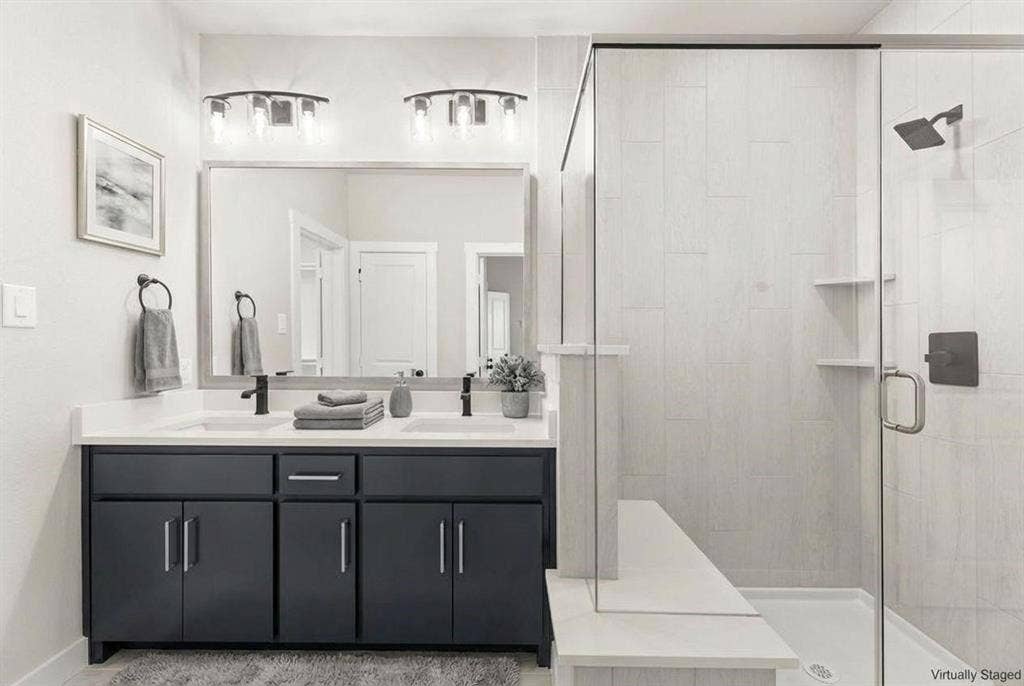 604 Nolan Lane, Wylie, TX 75098 Dual-sink vanity with dark cabinetry and white countertop, full-width mirror with dual overhead lighting, and walk-in shower featuring glass enclosure and stone-finish wall tile