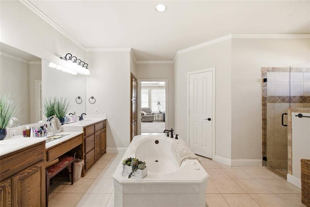 Jetted soaking tub with deck-mounted bronze faucet, walk-in shower with decorative tile inlay, arched window, and neutral tile flooring