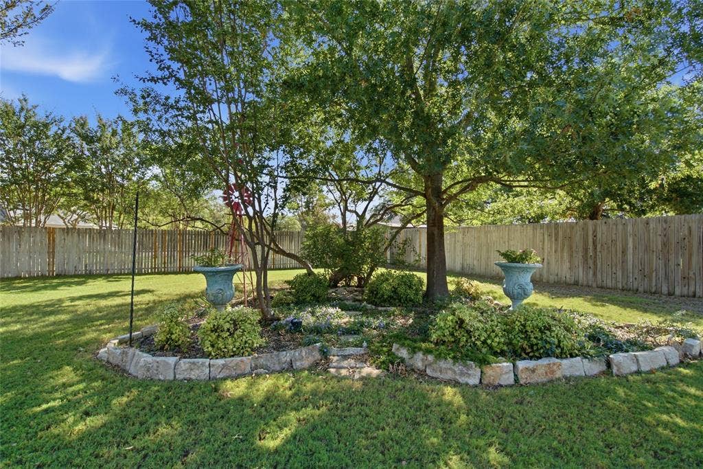 Backyard featuring a stone-edged garden bed, mature shade trees, decorative pedestal planters, and a full-perimeter wood fence