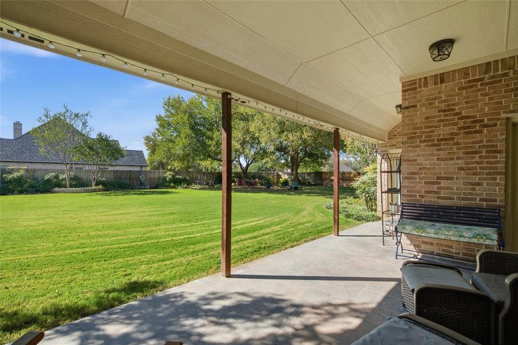 Covered patio with flagstone flooring, brick exterior wall, and expansive lawn with mature trees enclosed by wood fencing