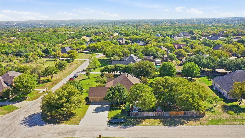Aerial perspective of a multi-gabled residence featuring an expansive lot, mature trees, a concrete driveway, and wooden perimeter fencing