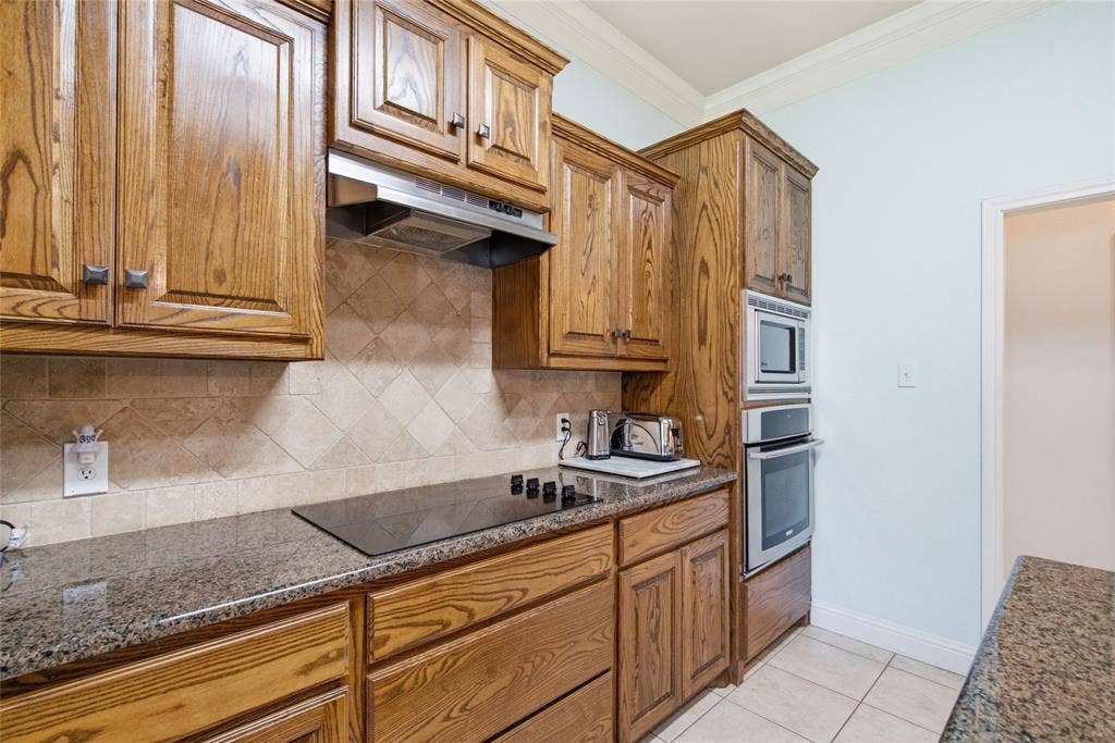 Kitchen featuring custom wood cabinetry, stone-surface countertops, tile backsplash, stainless steel cooktop with integrated range hood, and built-in wall oven and microwave