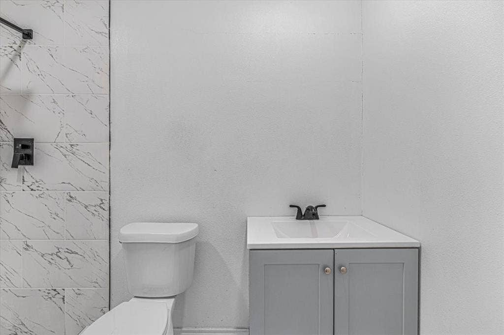 13521 Flagstone Lane, Dallas, TX 75240 White ceramic toilet, corner vanity with gray cabinetry, white countertop, and black faucet, alongside a shower with white marble-look tile and black fixtures