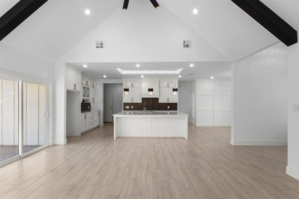 Vaulted ceilings with exposed beams, light wood-finish flooring, white kitchen cabinetry, kitchen island with seating, and sliding glass doors to the exterior