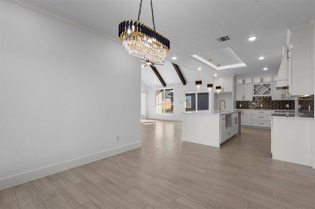 Wood-finish flooring, vaulted ceiling with exposed beams, pendant lighting, white cabinetry, and a kitchen island with integrated sink