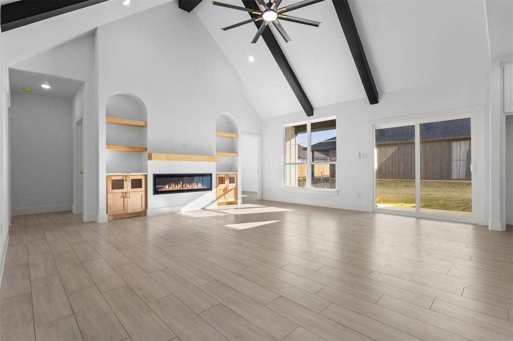Vaulted ceiling with exposed beams, wood-finish flooring, built-in shelving with lower cabinetry, linear electric fireplace, and sliding glass door to exterior