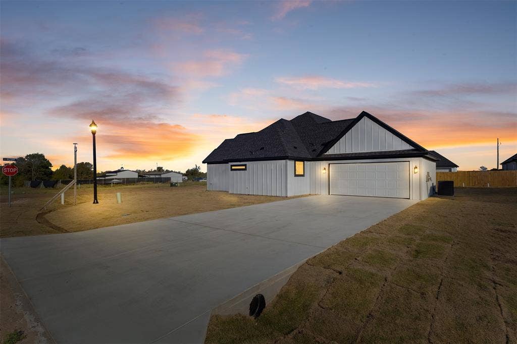 Board and batten siding, dark shingled roof, attached garage with decorative sconces, concrete driveway, and exterior lighting
