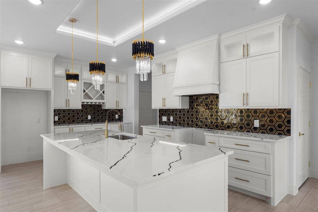 Kitchen featuring white cabinetry with gold-toned hardware, a spacious island with a veined countertop, a hexagon-patterned backsplash, and modern pendant lighting