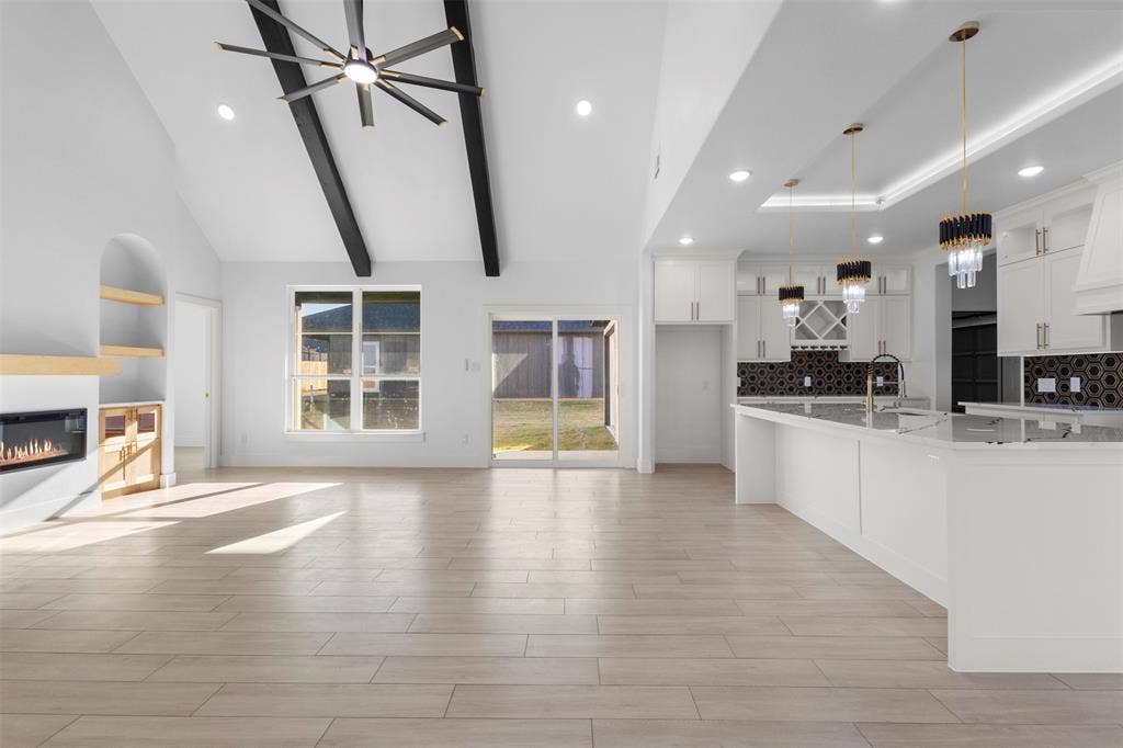 Vaulted ceilings with exposed beams, wood-finish flooring, built-in fireplace, and a kitchen featuring white cabinetry, an island, and decorative pendant lighting