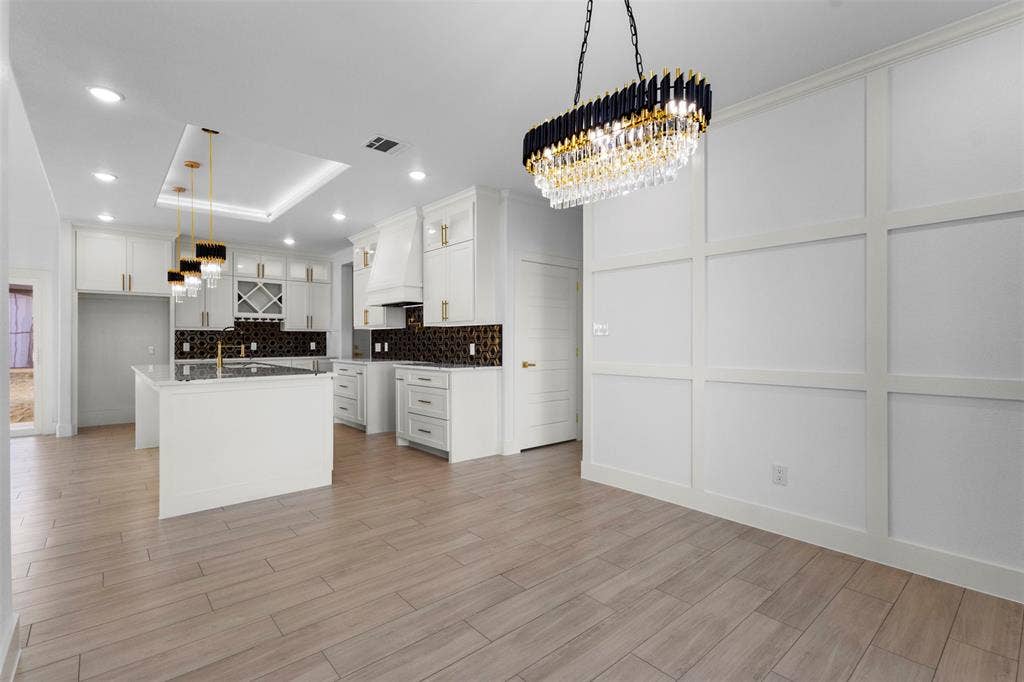 White shaker cabinetry, patterned tile backsplash, kitchen island with built-in sink, wood-finish flooring, and a decorative paneled wall