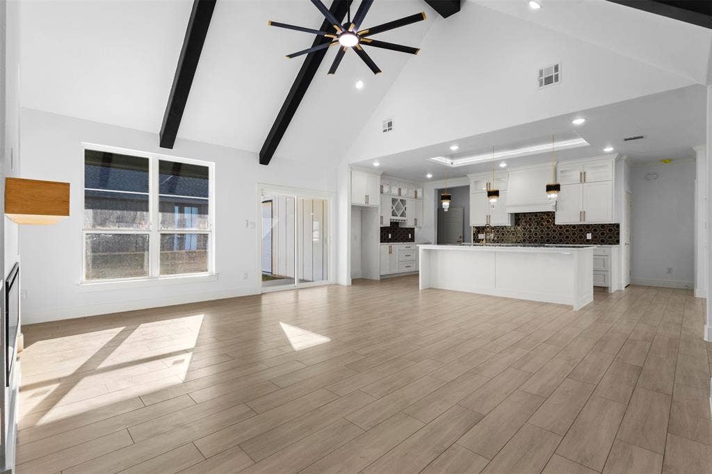 Vaulted ceiling with exposed beams, wood-finish flooring, white cabinetry, kitchen island with built-in sink, and patterned backsplash