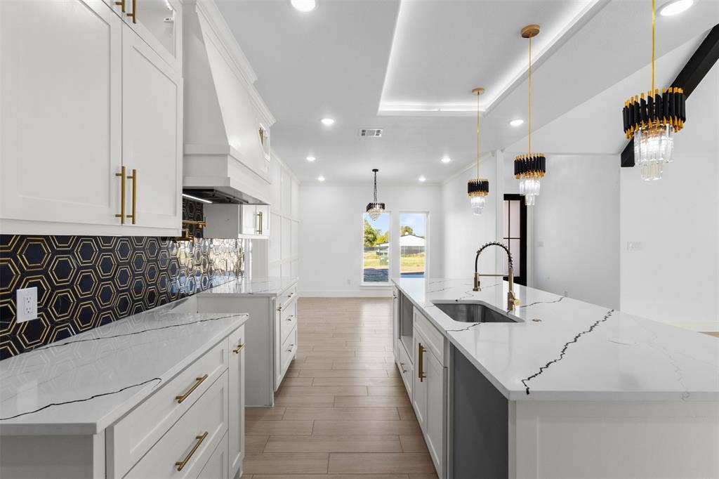 White shaker-style cabinetry, hexagon tile backsplash with gold accents, white countertops with prominent veining, kitchen island with integrated sink, and pendant lighting